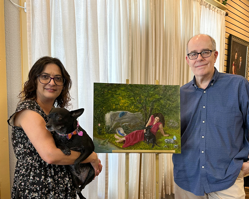 Client Michelle holding her dog Miya while standing beside artist Hayward Gaude and the finished fantasy portrait inside the studio.